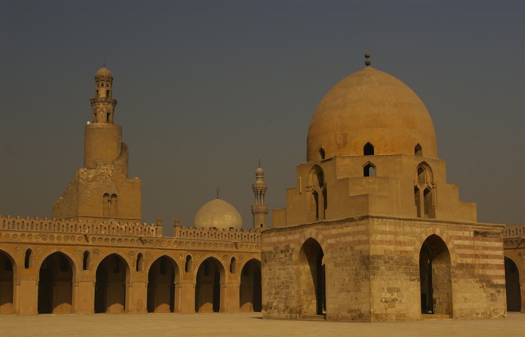Cairos ancient mosque courtyard with arched walls and large domes under a hazy sky.
