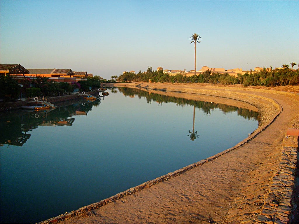 El Gounas tranquil canal scene with palm trees and distant buildings under a clear sky.