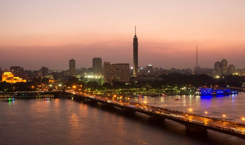 City skyline at dusk with a river, a lit-up bridge, like cruising the Nile in late summer.