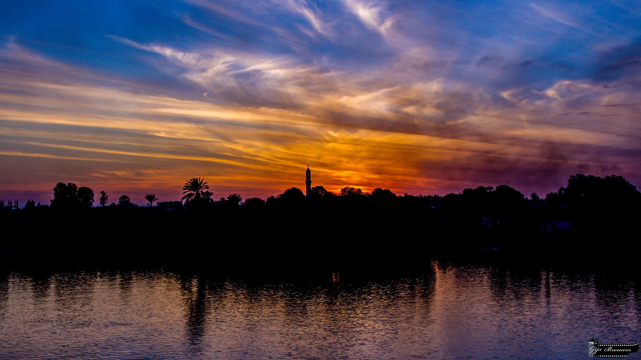 Nile sunset over a lake with silhouetted trees and wispy clouds.
