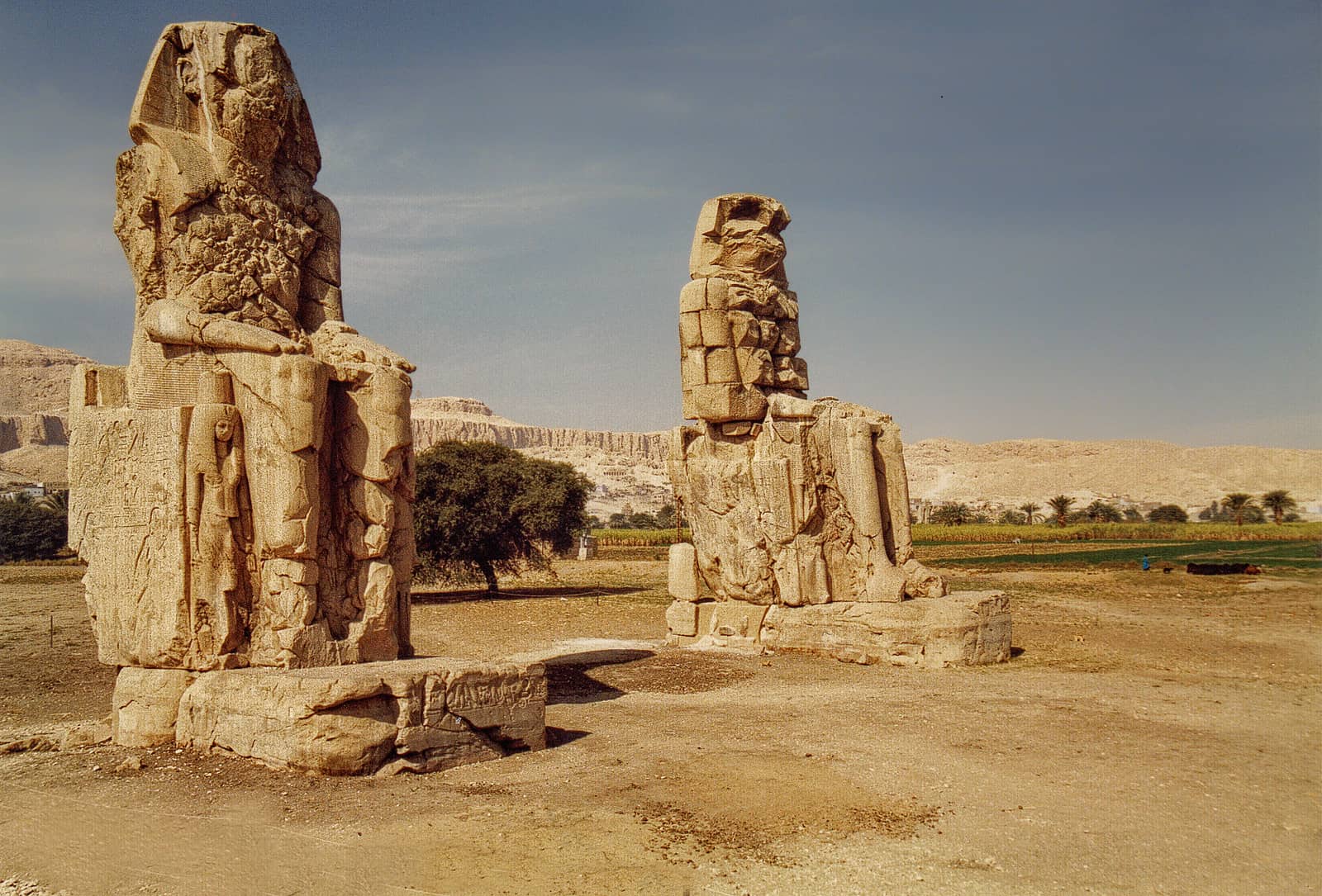 Two ancient stone statues stand near the Nile in a desert landscape under a clear sky.