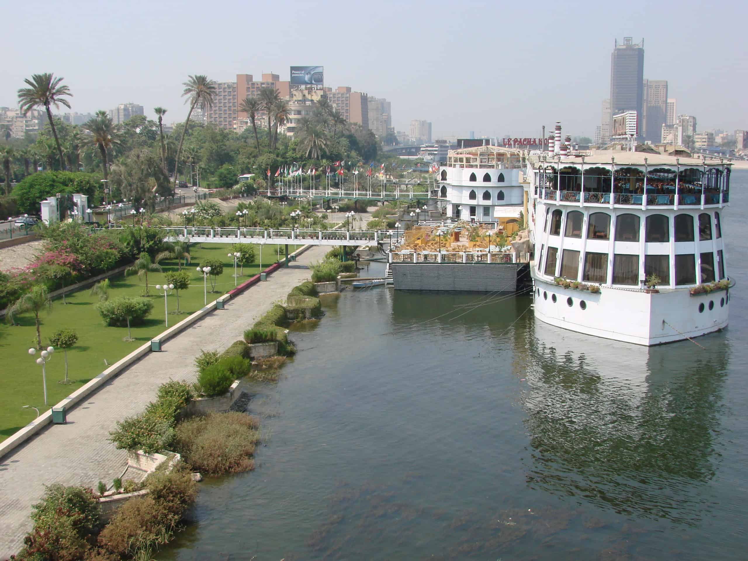 Large white ship docked by a lush riverside park with an Egypt-inspired cityscape behind.