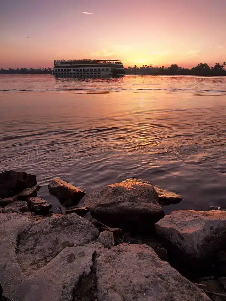 Boat on a river at sunset with rocky shoreline in the foreground.