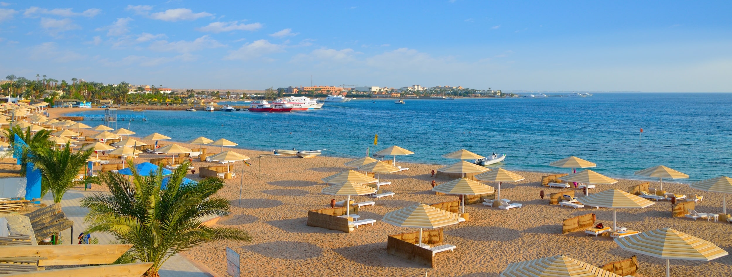 Sandy beach near Sharm El Sheikh Hotels, parasols by the blue sea; boats visible afar.