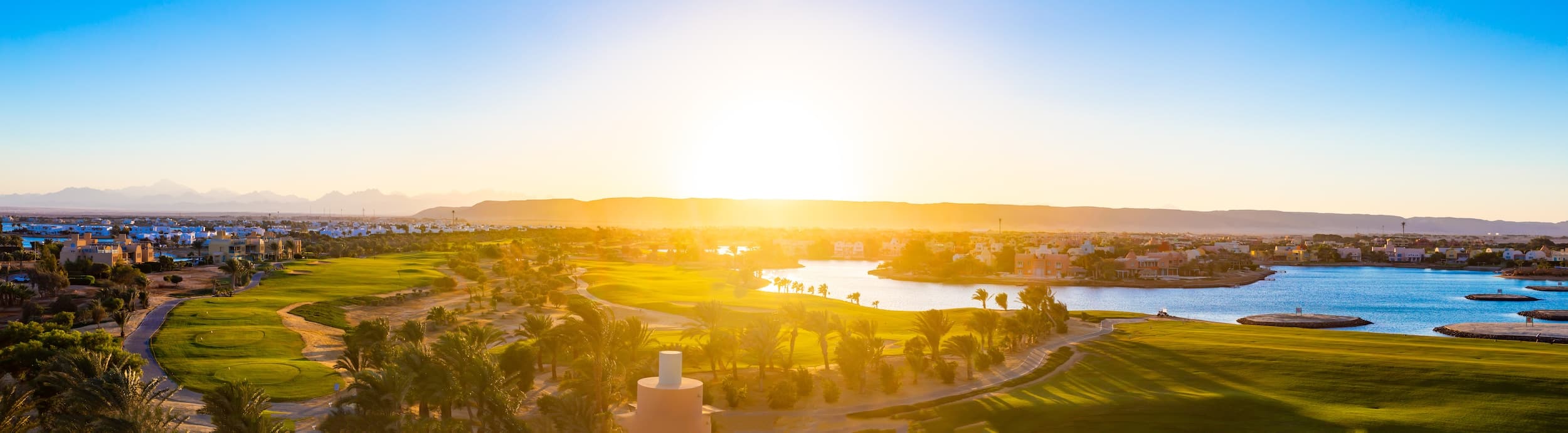 Sunset over a golf course with a lake, palm trees, and El Gouna Hotels in the distant mountains.