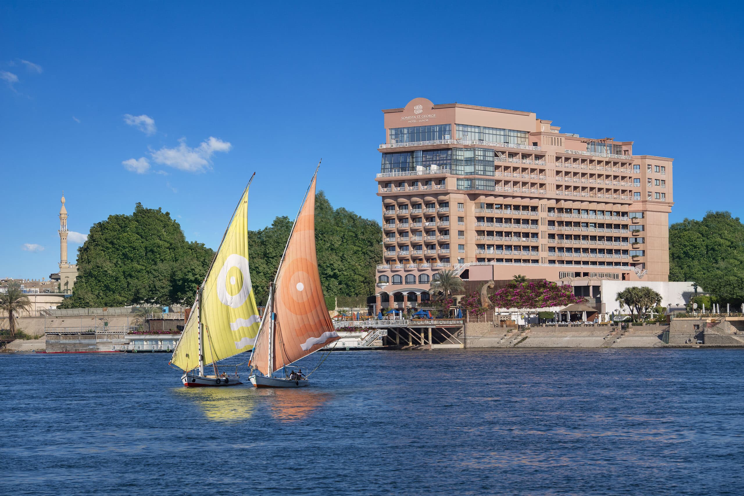 Sailboats with colorful sails glide by green trees near Sonesta St George Luxor on the river.