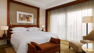 Room at Sheraton Cairo Hotel & Casino with a made bed, two lamps, artwork, chair, and sheer curtains.