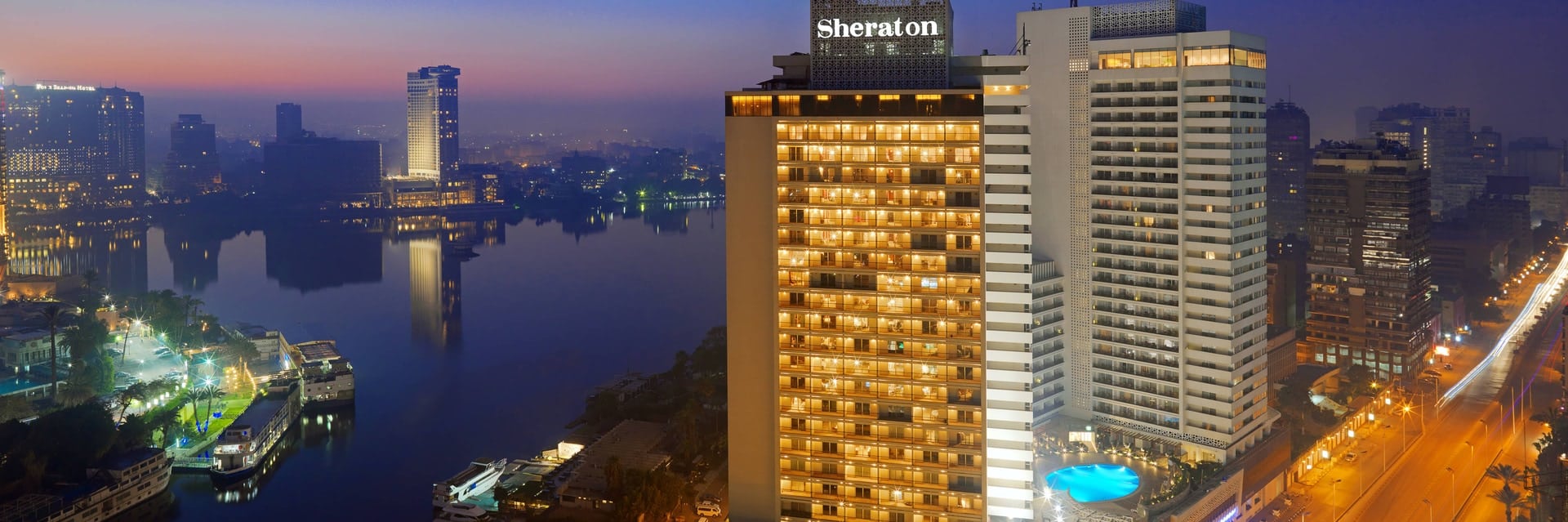 Night view of Sheraton Cairo Hotel & Casino by a river, city lights glowing in the background.