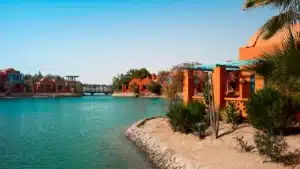 Orange buildings by a turquoise lake, resembling Sheraton Miramar Resort, with a wooden bridge afar.