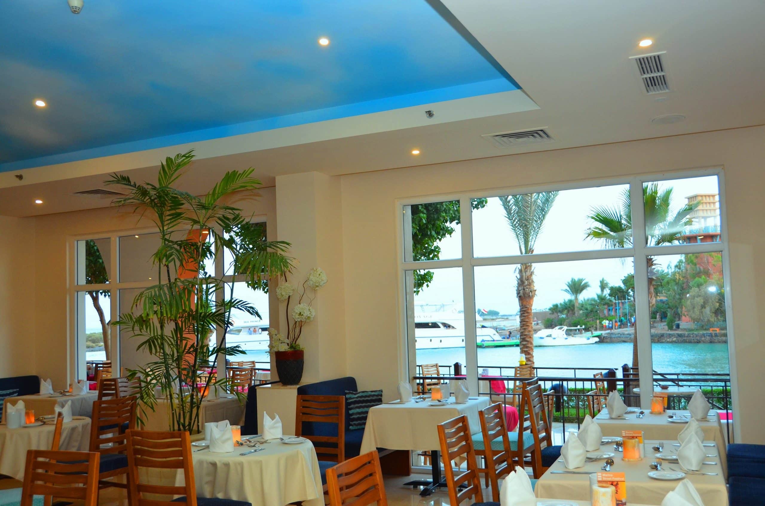 Three Corners Ocean View restaurant with set tables and large windows framing boats and palm trees.
