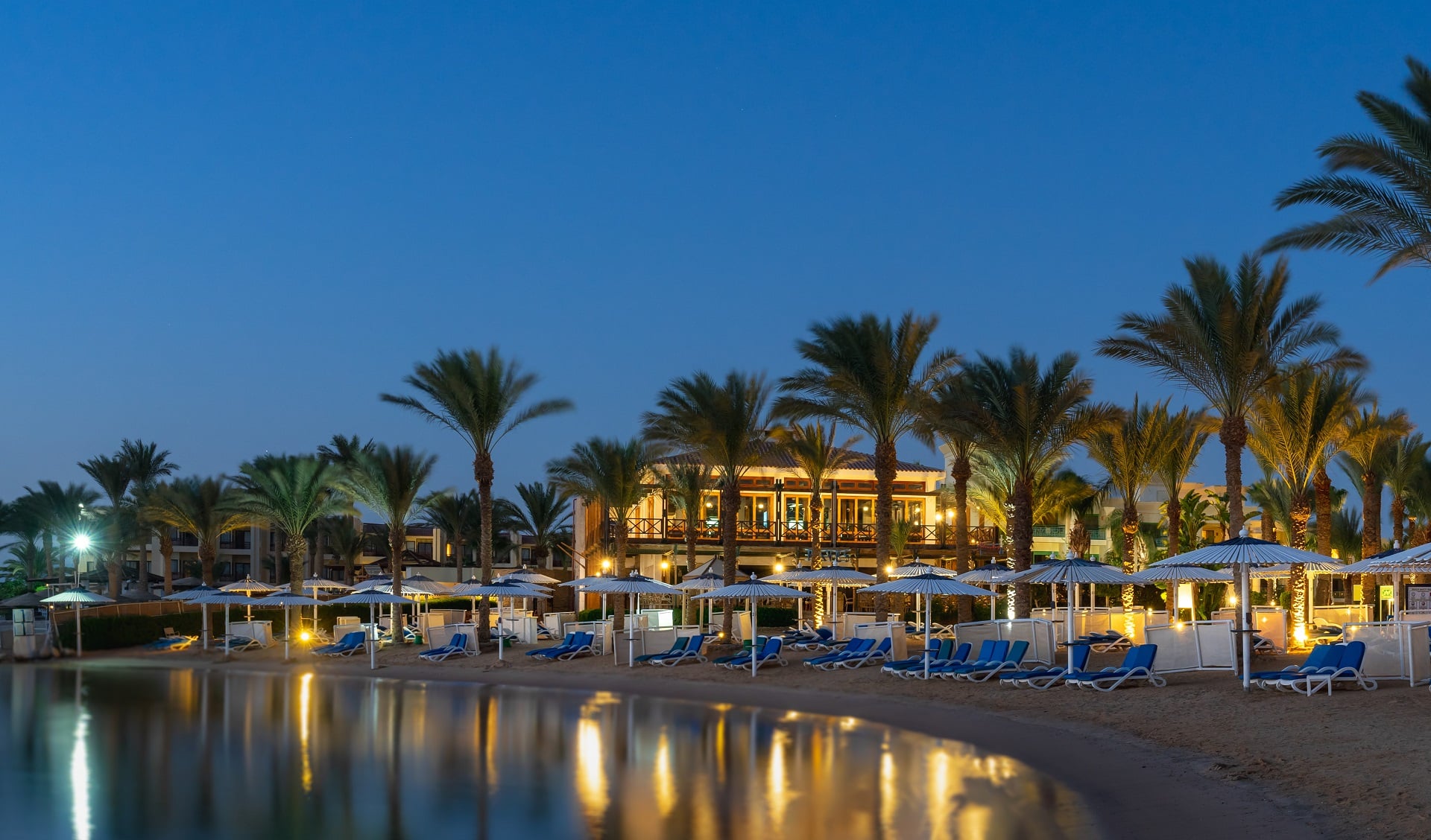 Swiss Inn Resort Hurghada: Beachfront oasis with palm trees, sun loungers, and lit buildings at dusk.