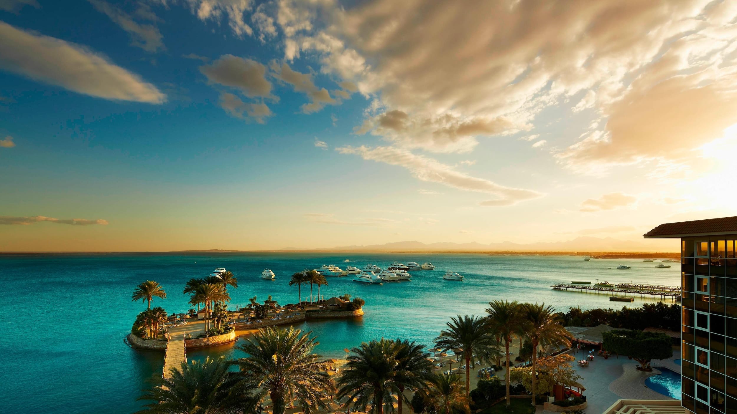 Scenic view of Hurghada Marriott Beach Resorts coast with palm trees, boats, and a sunny dusk sky.