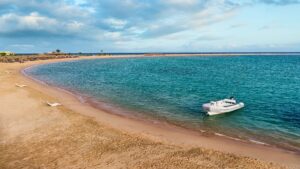 A small white boat on Sheraton Soma Bays sandy beach, with clear blue water and a cloudy sky.