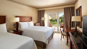 Room with two beds, desk, chairs; patio opens to garden seating at Cairo Marriott Hotel.