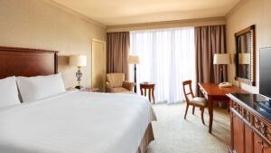 Room at Cairo Marriott Hotel with bed, desk, armchair, lamps, TV by a large window.