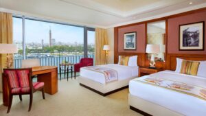 The Nile Ritz-Carlton room with two beds, a desk, and a city view through a large window.