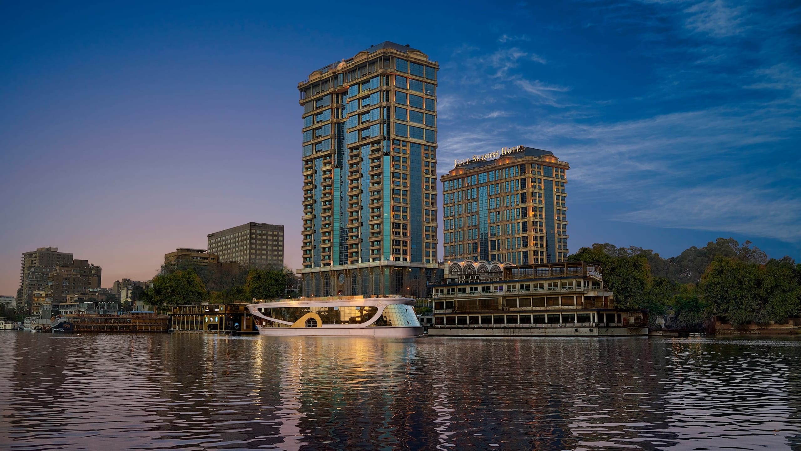Two tall buildings by a river at sunset, with Four Seasons at The First Residence in view.